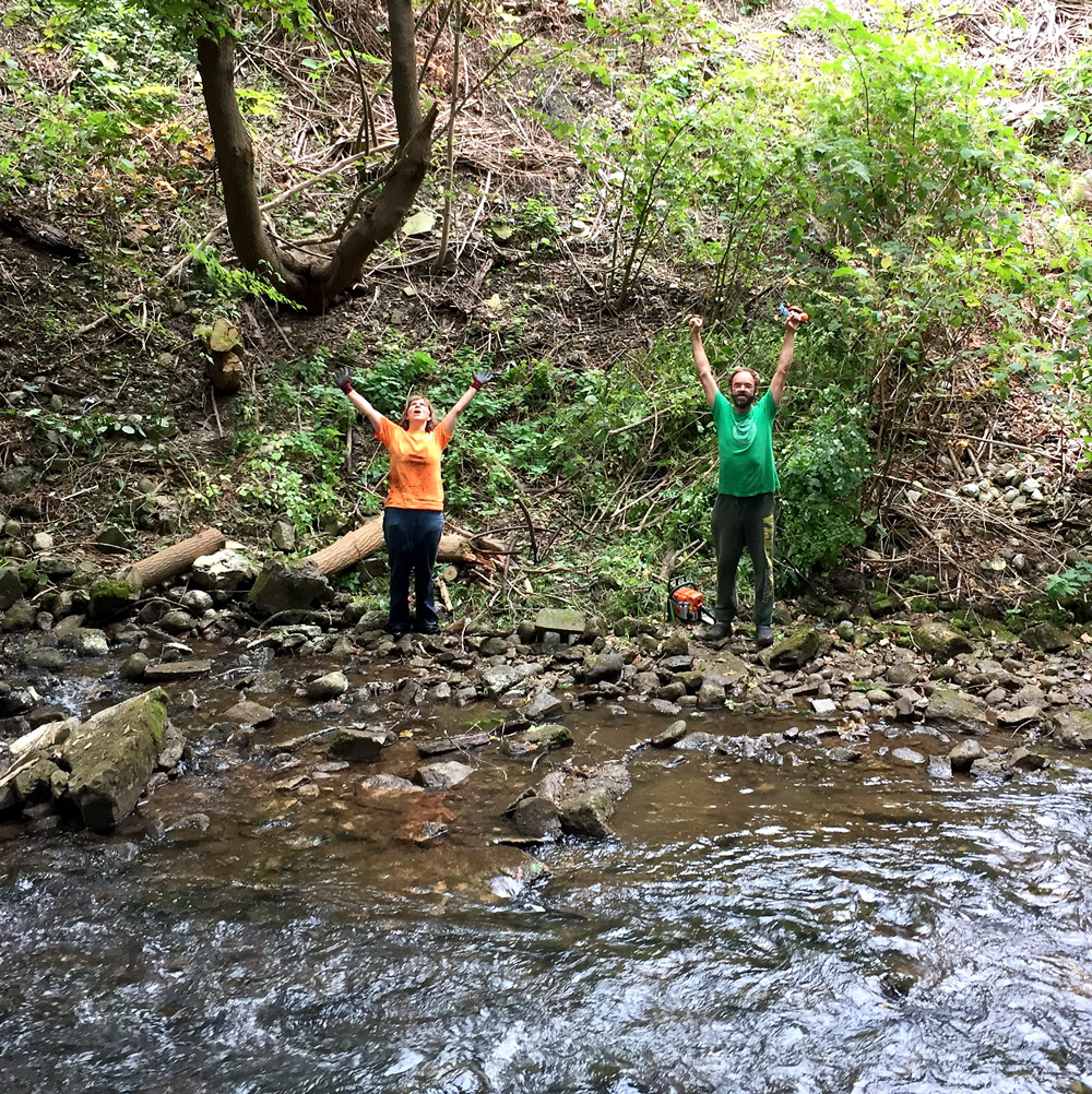 2 volunteers raise their hands in joy during a clean up day by the river